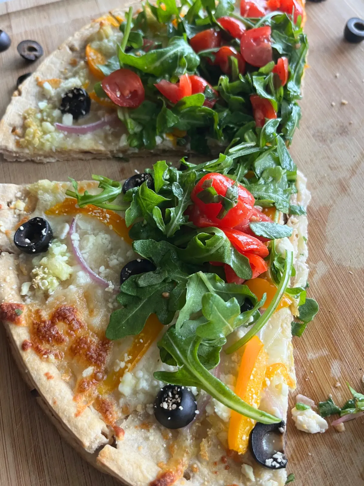 A close-up of two slices of Mediterranean diet pizza on a wooden cutting board after slicing.