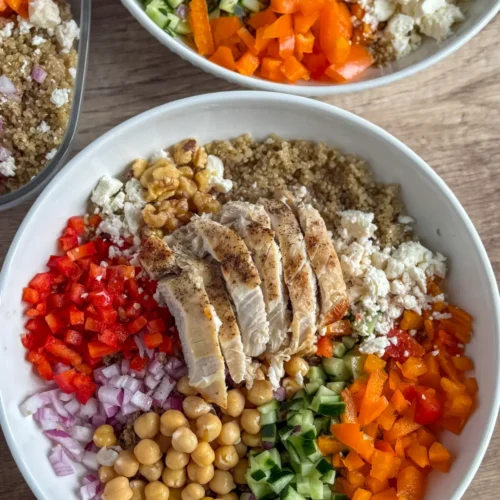 Two fully assembled Mediterranean grain bowls on a counter next to a meal prep container of a bowl.