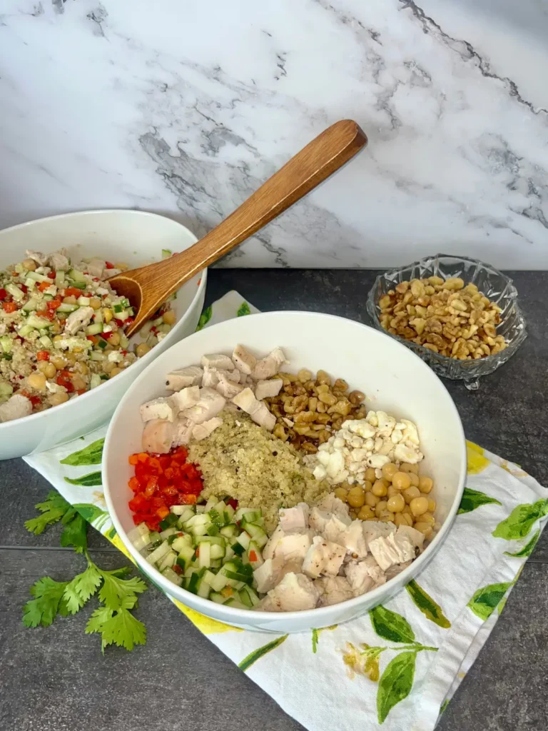 A Mediterranean quinoa bowl on a towel, next to a larger bowl and a bowl of chopped walnuts.