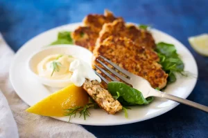 Salmon patties on a plate with tartar sauce, a lemon wedge, and a fork.