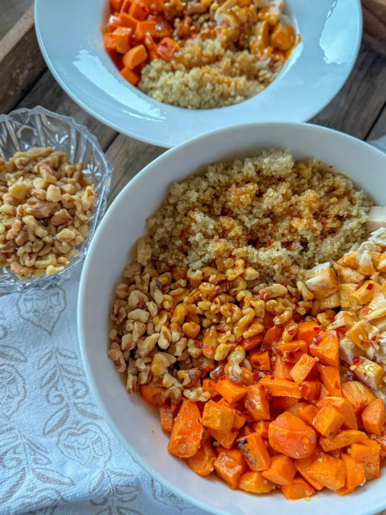 A large bowl of sweet potatoes, quinoa, chopped walnuts, and chicken next to a small bowl of it on a wooden board.