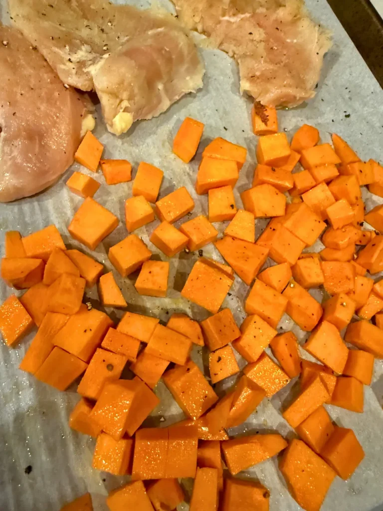 Cubed sweet potatoes on a baking sheet lined with parchment next raw chicken breasts.