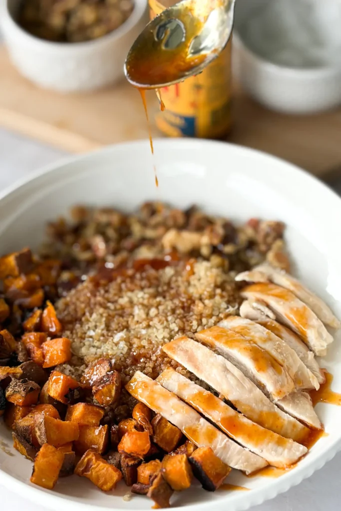 A spoon drizzling the honey sriracha sauce onto a fully assembled sweet potato chicken and quinoa bowl.