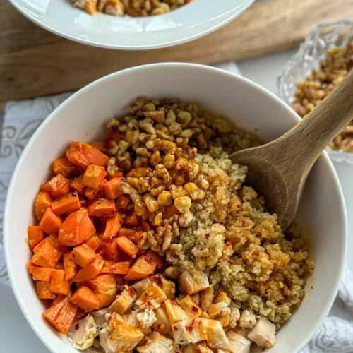 A large bowl with roasted sweet potatoes, chopped walnuts, quinoa, and chicken drizzled with honey sriracha sauce on a counter.