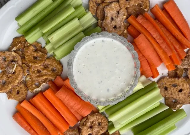 Healthy ranch dip on white platter surrounded by cut carrots and celery.