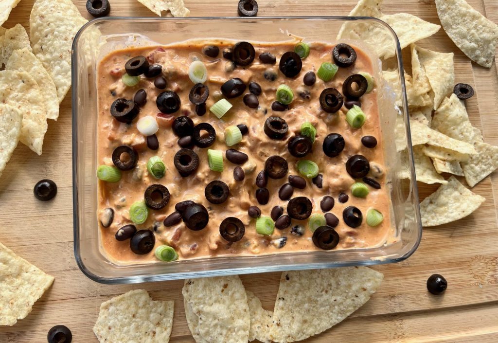 A high protein taco dip topped with olives and green onions in a glass container on a cutting board surrounded by chips.