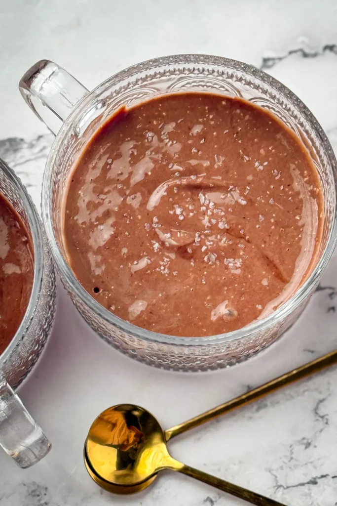 A bowl of dark chocolate pudding sprinkled with sea salt next to a gold spoon.