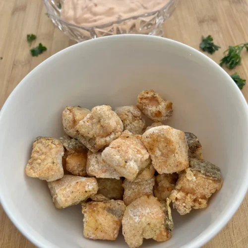 Crispy salmon bites in a white bowl on a wood cutting board next to a bowl of bang bang sauce.