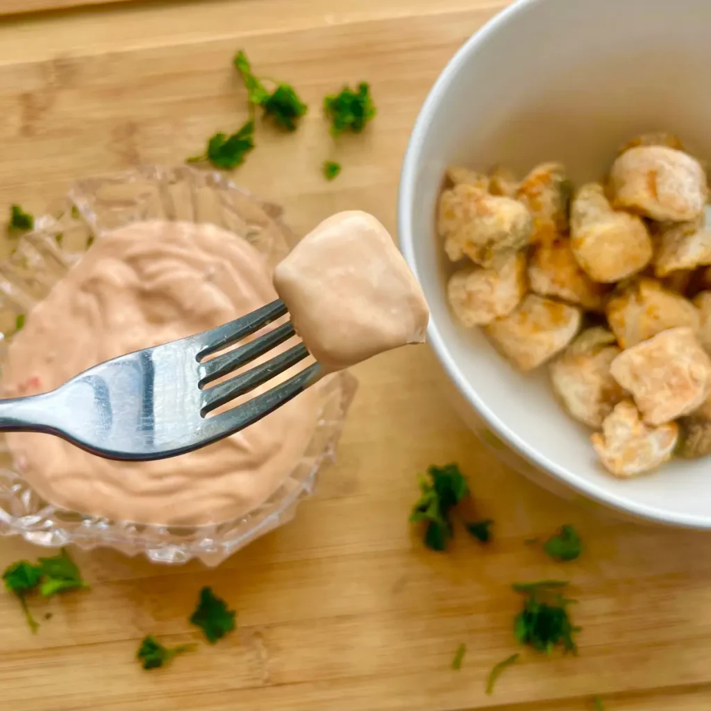 A close-up of a crispy salmon bites on a fork with a bowl of salmon bites and bang bang sauce in the background.