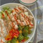 A birds-eye view of a crispy tofu, rice, and mixed vegetable bowl on a table next to a bowl of Greek yogurt bang bang sauce.