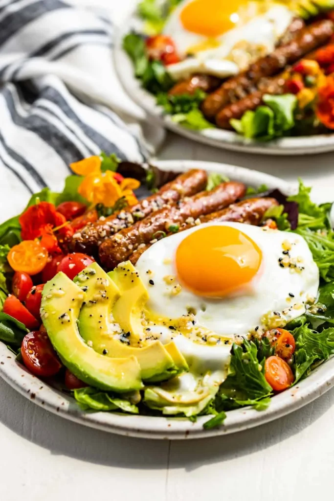 Two plates of dairy-free, gluten-free breakfast salad topped with eggs, sausage, avocado and veggies.