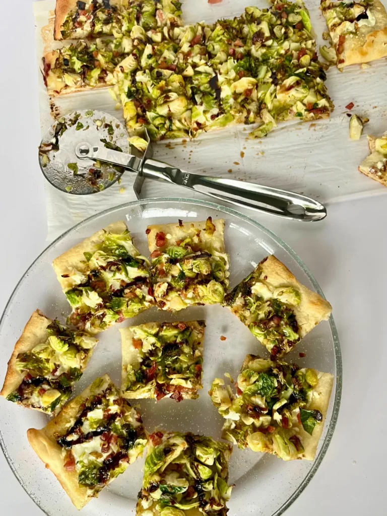 Goat cheese and Brussel sprout pizza slices on a platter next to a cutting board with more pieces on it. 