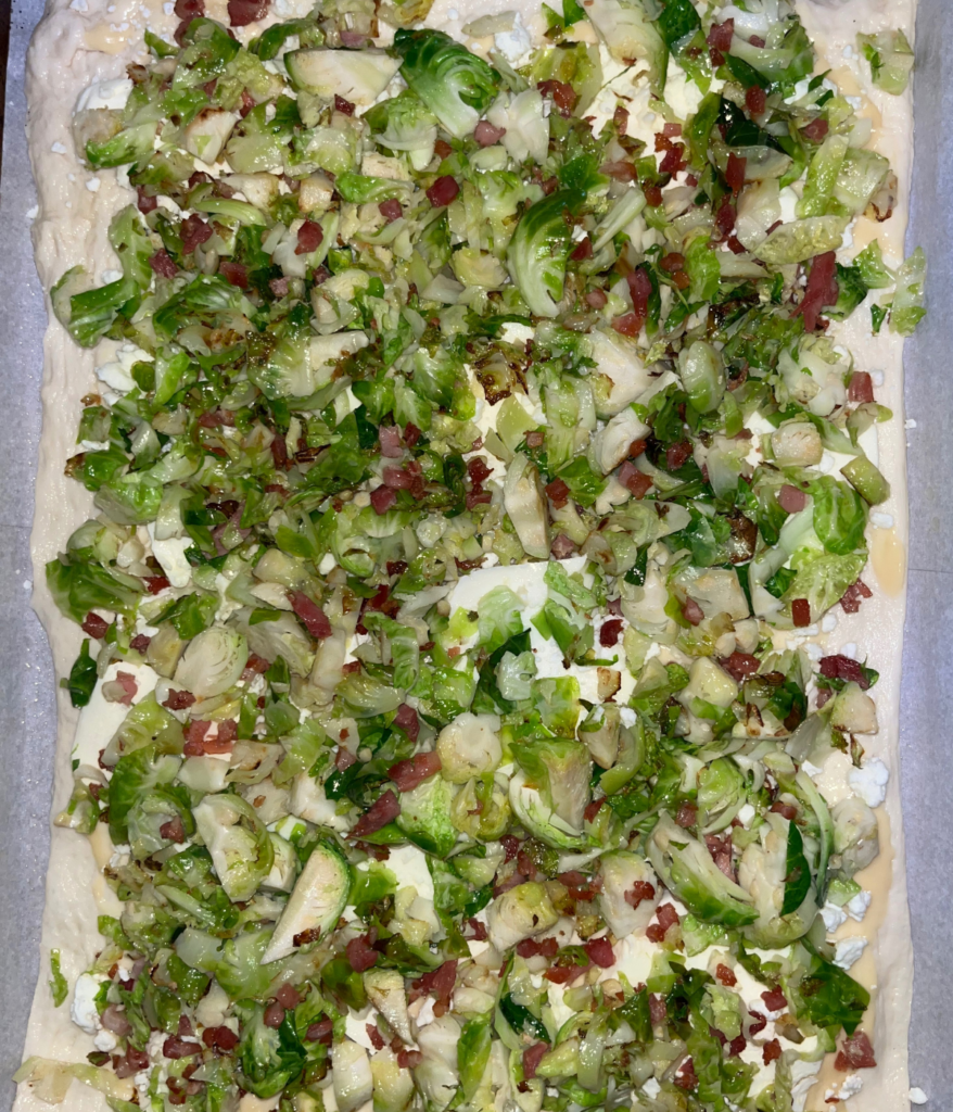 Raw pizza dough topped with mozzarella cheese, goat cheese, crispy pancetta, and Brussel sprouts before baking in the oven.