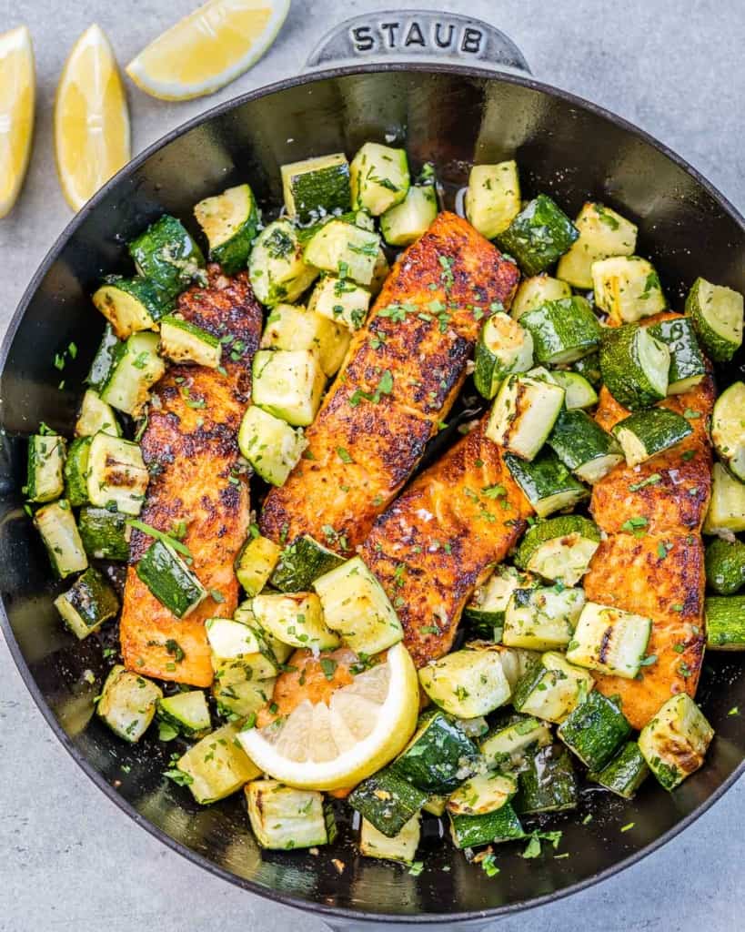 A birds-eye view of cajun baked salmon with zucchini in a skillet with garnished lemon slices.