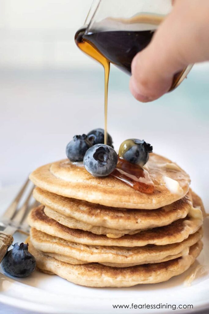A stack of gluten-free pancakes topped with blueberries and someone pouring maple syrup on them.