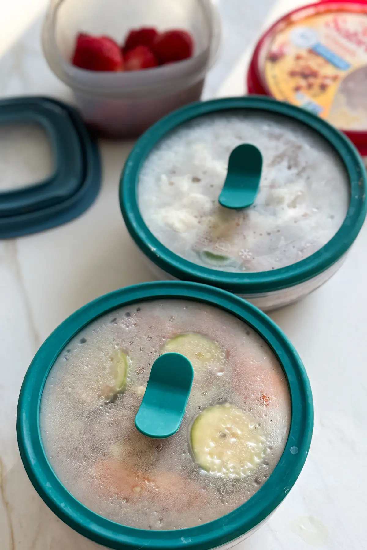 Two Anyday storage containers with lids on a counter next to a carton of hummus and a storage container with cut up strawberries.
