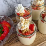 Four mini strawberry cheesecake protein cups on a wooden cutting board next to a bowl of strawberries.