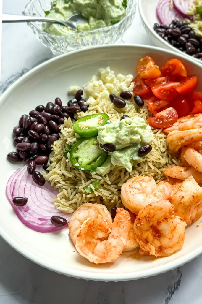 A fully assembled shrimp taco bowl topped with the avocado yogurt sauce and jalapeno slices next to another bowl.