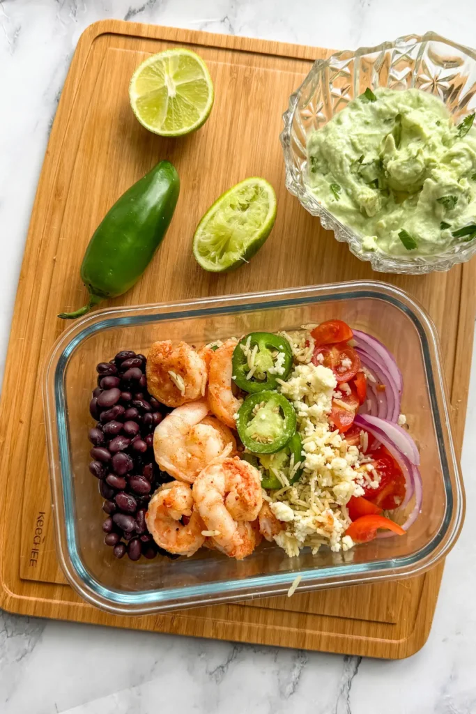 A birds-eye view of a shrimp and rice meal prepped taco bowl on a wooden cutting board next to the avocado yogurt sauce and a lime.