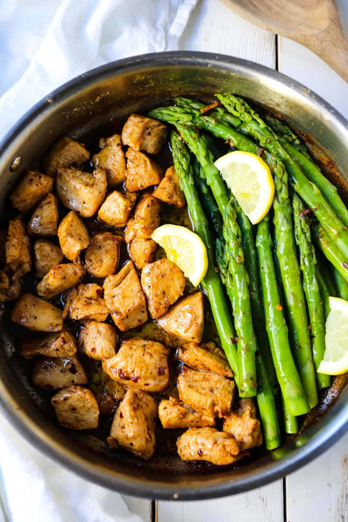  birds-eye view of garlic butter chicken in a stainless steel pan with asparagus on a table.