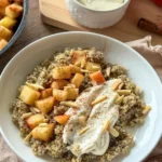 An apple cinnamon quinoa bowl topped with the Greek yogurt and cream cheese mixture in a white bowl on a table next to more yogurt and an apple.