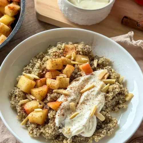 An apple cinnamon quinoa bowl topped with the Greek yogurt and cream cheese mixture in a white bowl on a table next to more yogurt and an apple.