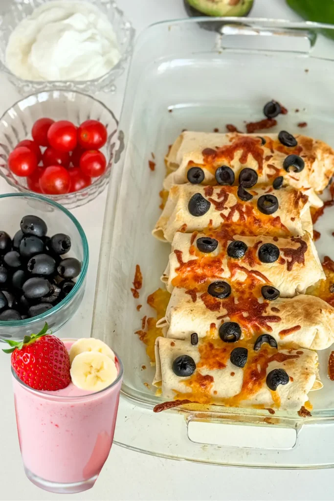 Breakfast burritos in a baking dish next to a strawberry smoothie, a bowl of tomatoes, a bowl of olives, and an avocado.