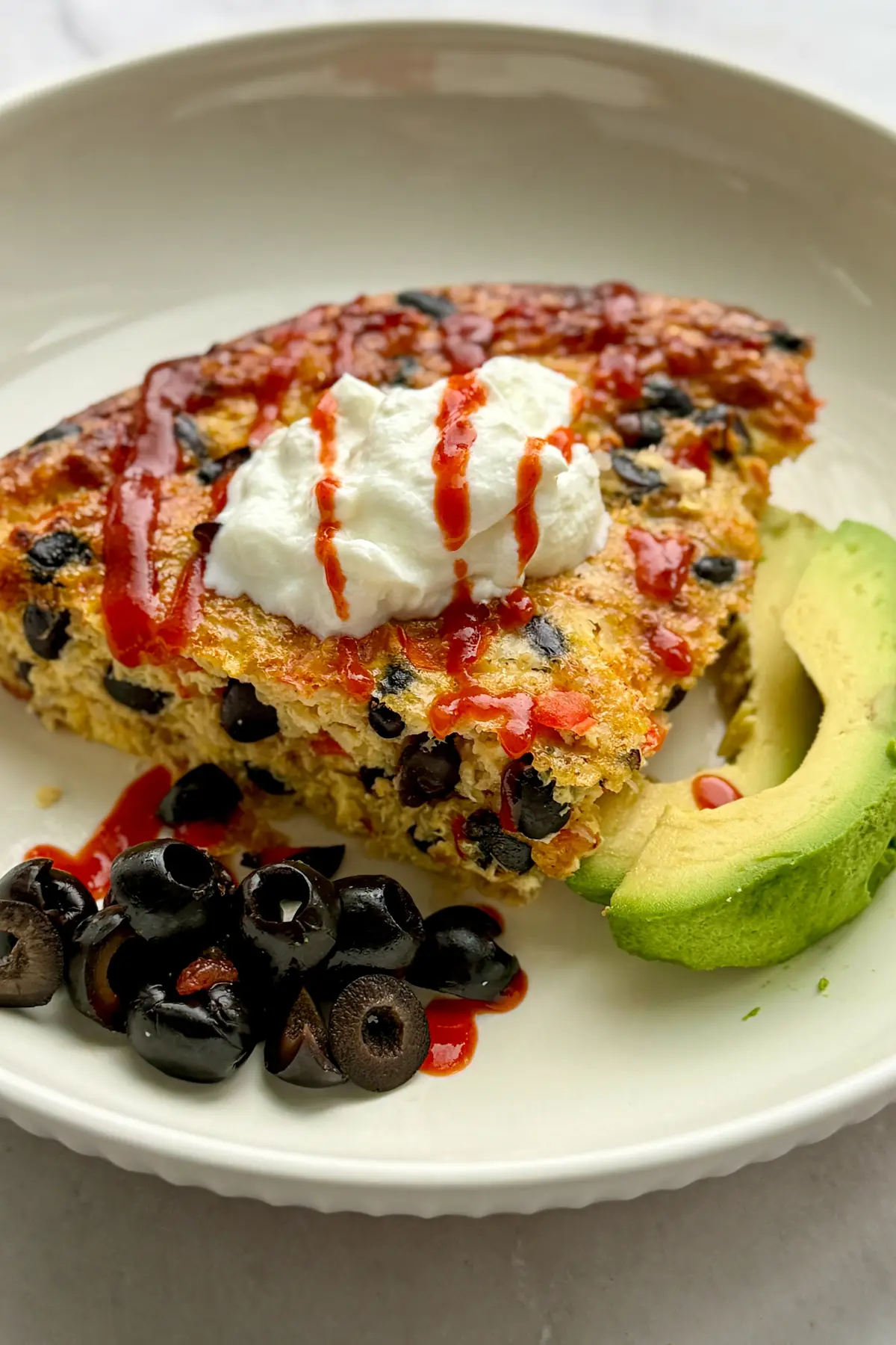 A slice of quinoa breakfast baked topped with Greek yogurt and hot sauce on a plate next to avocado and black olives.