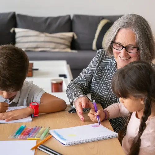 A grandmother helping two of her grandchildren color at a table.