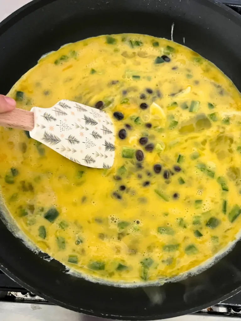 Eggs, black beans, and jalapenos cooking in a pan - being stirred by a spatula.