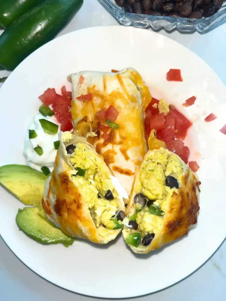 A burrito cut in half on a plate next to avocado slices, tomatoes, and Greek yogurt.