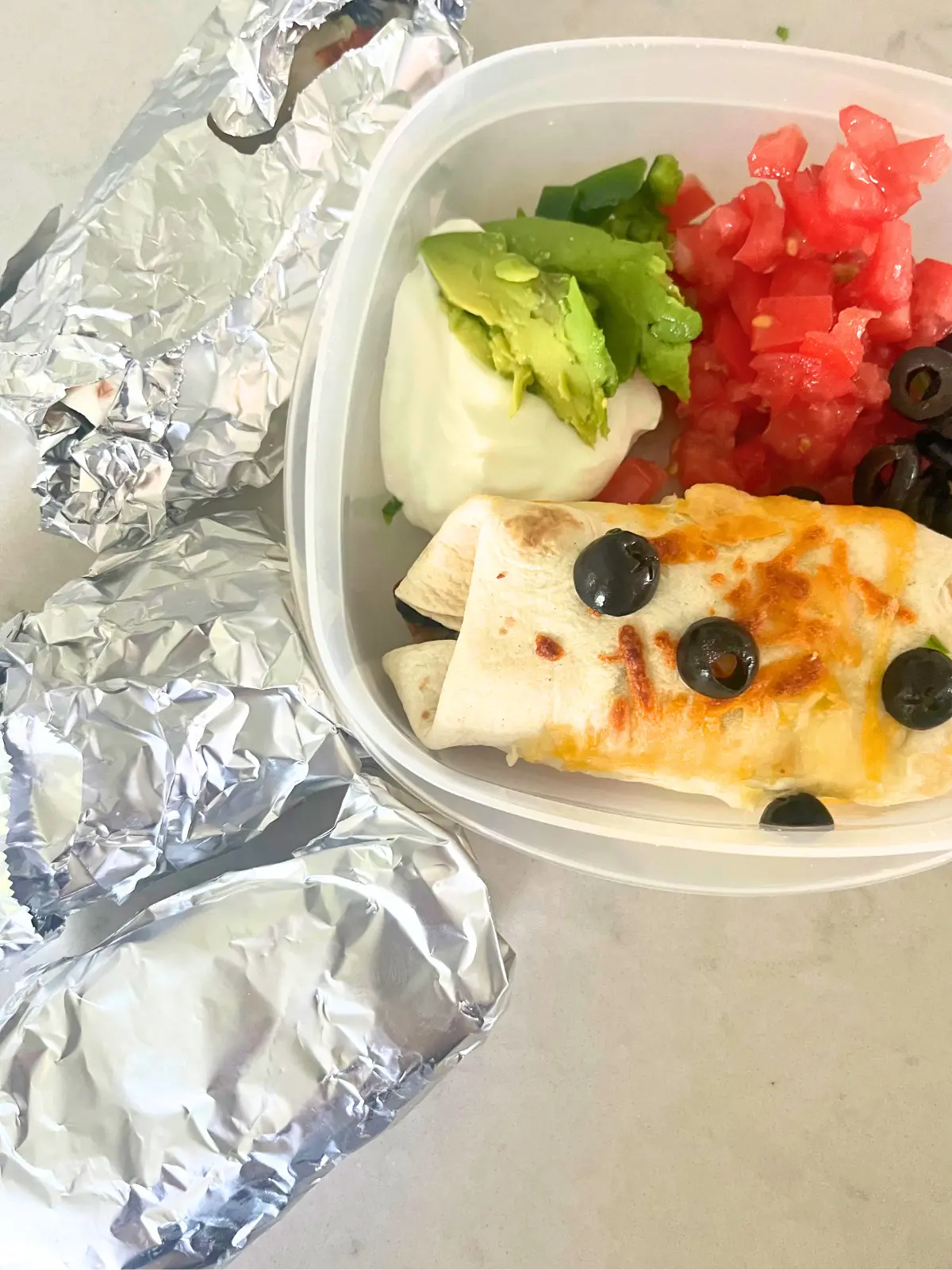 Burritos wrapped in tinfoil next to a storage container with a breakfast burrito and toppings.