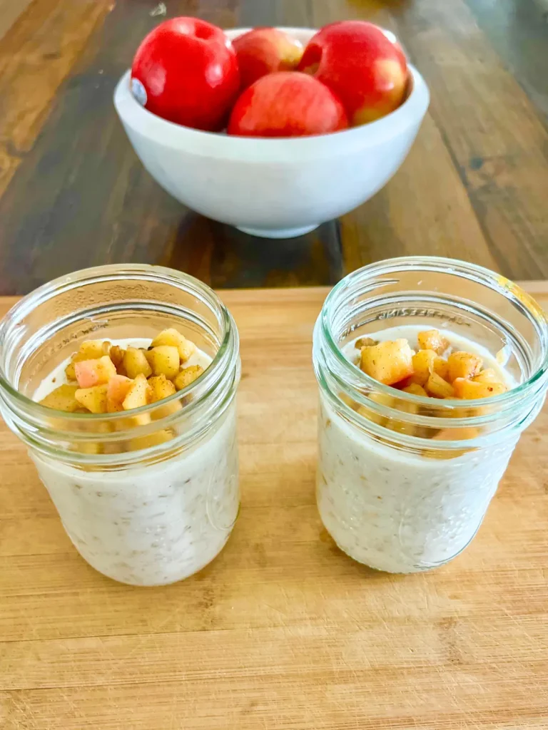Two mason jars filled with caramelized apples overnight oats on a wood cutting board on a dining room table with a bowl of apples in the background.