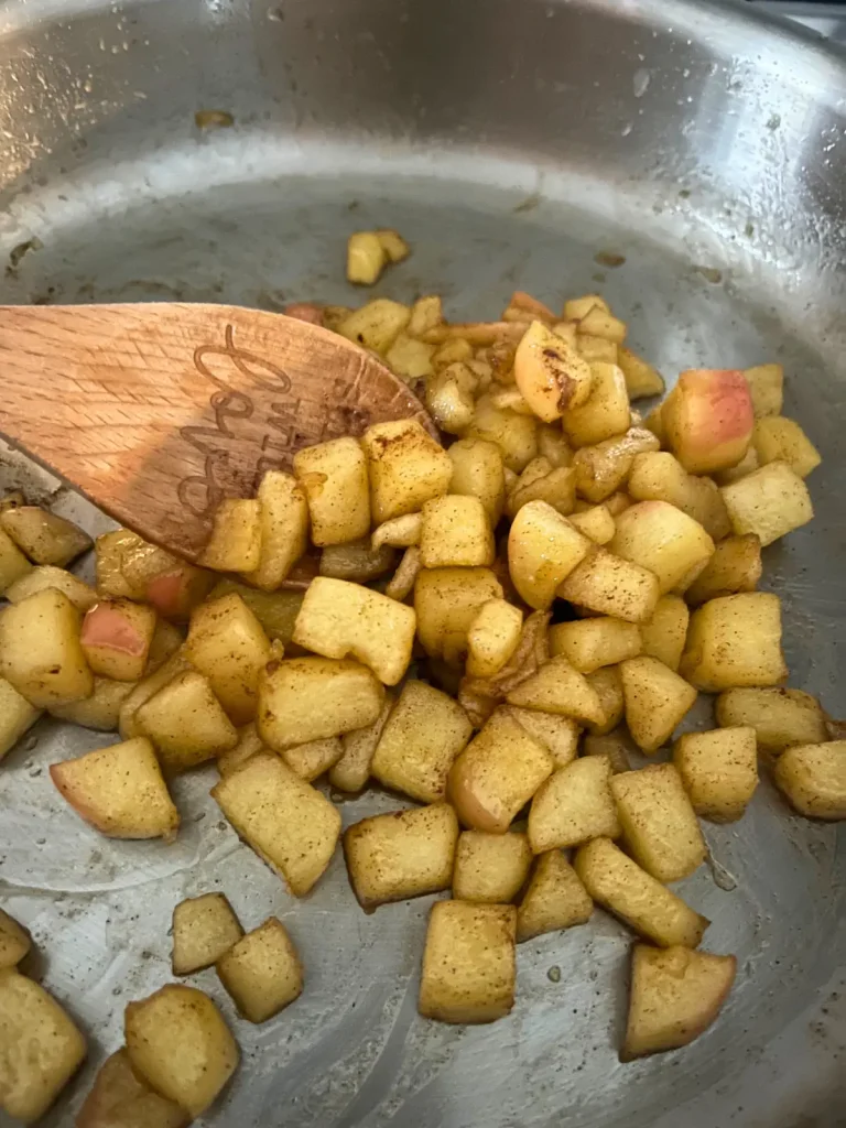 Caramelized apples in a stainless steel pan after cooking, being stirred with a wooden spoon.