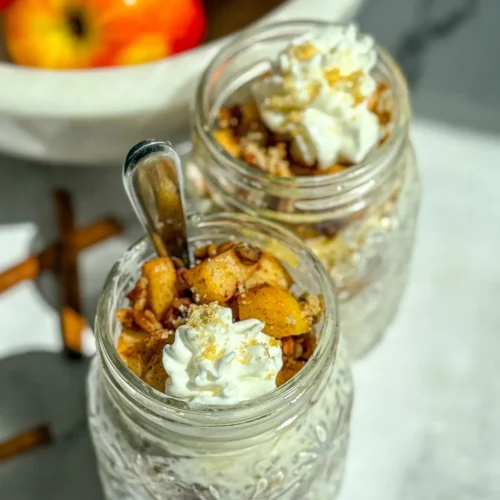 A birds-eye view of two apple pie overnight oats with pecans and whipped cream on top on a counter next to a bowl of apples.