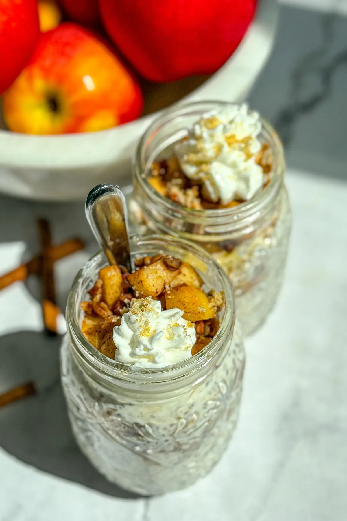 A birds-eye view of two apple pie overnight oats with pecans and whipped cream on top on a counter next to a bowl of apples.