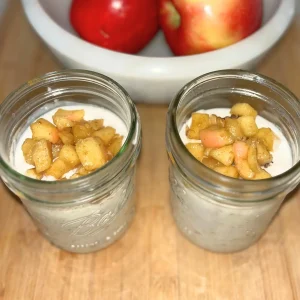 Two glass jars filled with overnight oats topped with caramelized apples and chopped walnuts on a wood cutting board next to a bowl of apples.