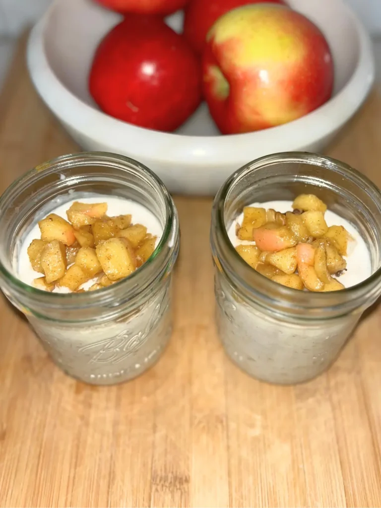 Two glass jars filled with overnight oats topped with caramelized apples and chopped walnuts on a wood cutting board next to a bowl of apples.