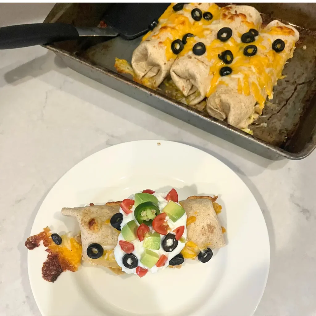 A burrito on a white plate next to a pan of the rest of the burritos.