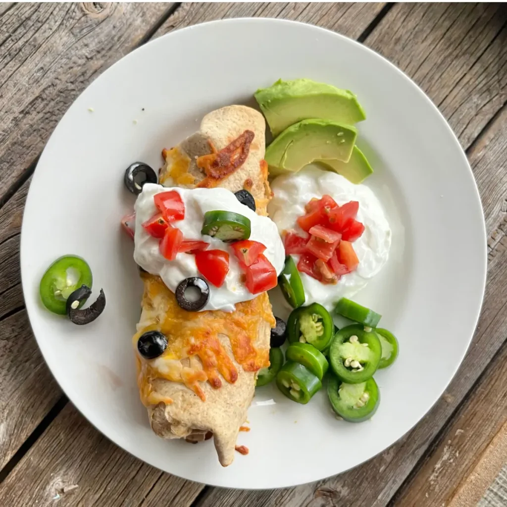 A high protein breakfast burrito with a crispy cheese crust on a white plate topped with Greek yogurt, sliced avocado, and diced tomatoes.