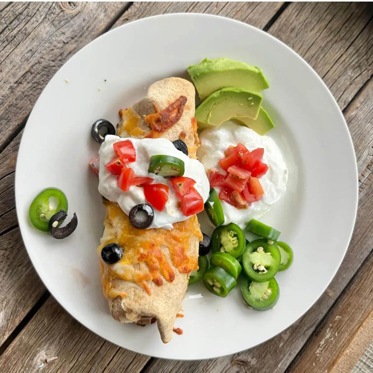 A high protein breakfast burrito with a crispy cheese crust on a white plate topped with Greek yogurt, sliced avocado, and diced tomatoes.