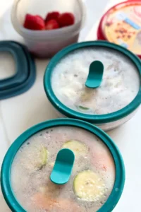 Two storage containers on a counter next to a bowl of strawberries and a carton of hummus.