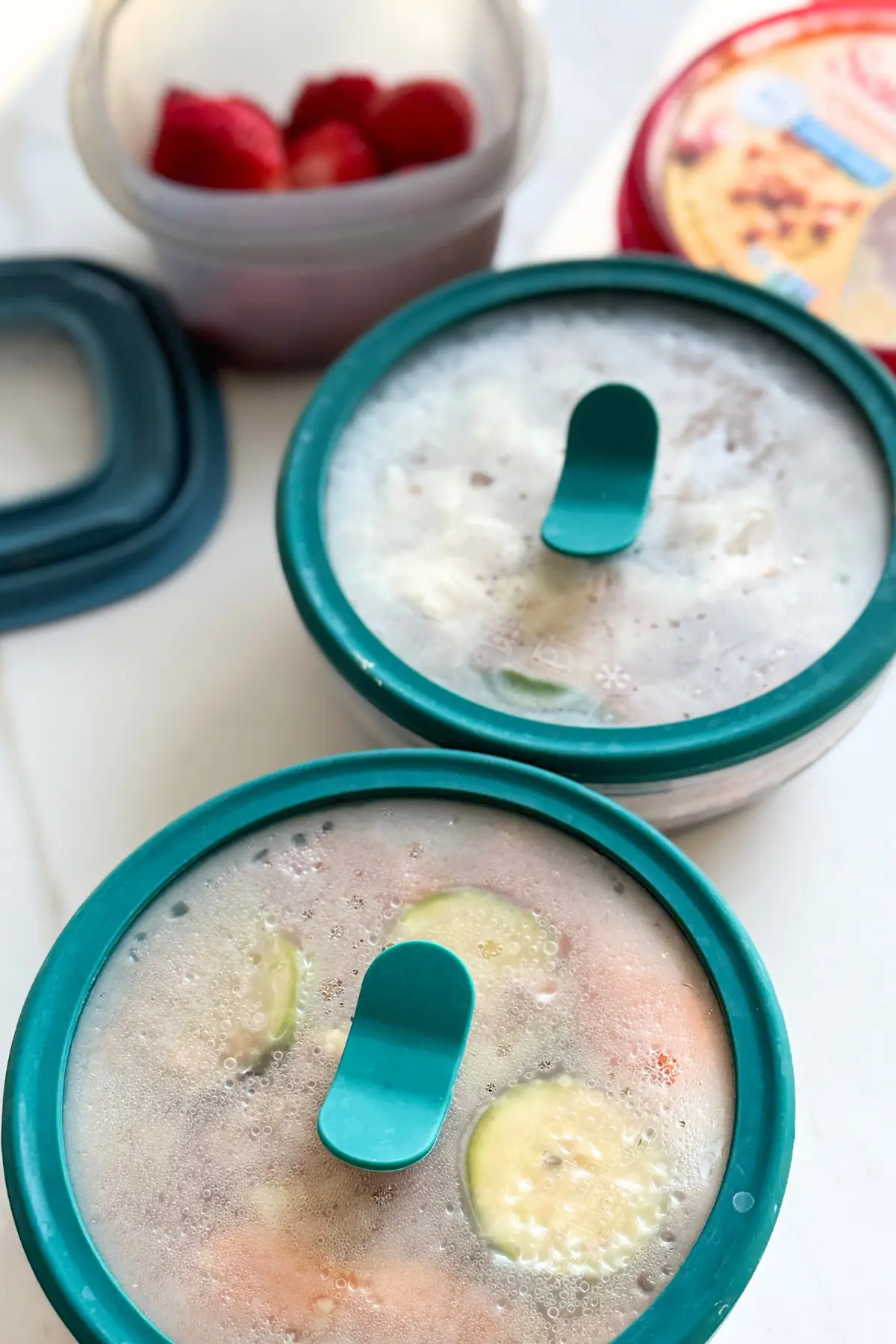 Two storage containers on a counter next to a bowl of strawberries and a carton of hummus.