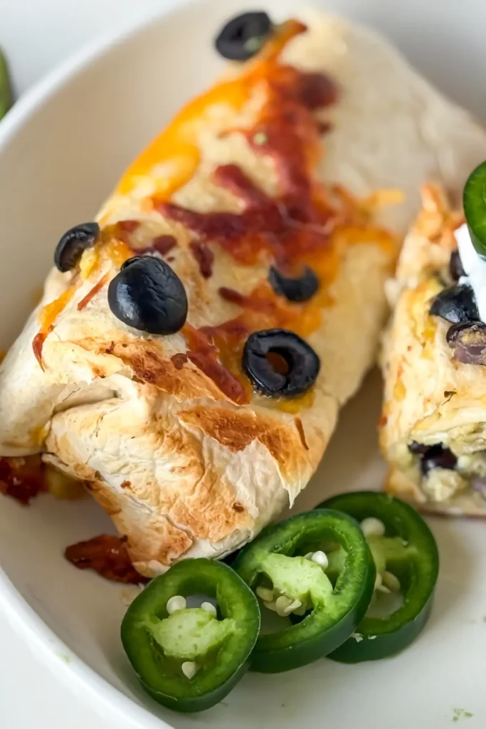 A crispy breakfast burrito on a plate next to a halve of a burrito garnished with olives and jalapenos.