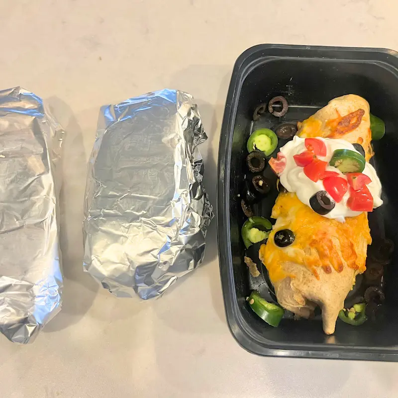 Two breakfast burritos wrapped in foil, and one in a storage container topped with Greek yogurt, diced tomatoes and jalapenos.