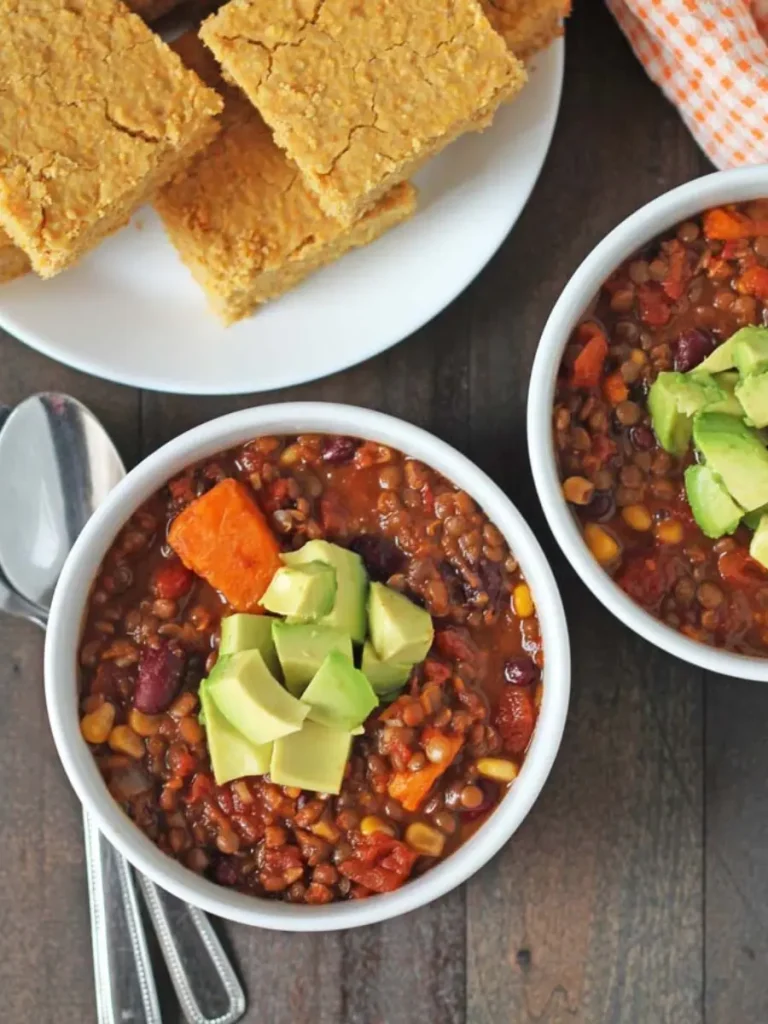 Two bowls of lentil sweet potato chili topped with avocado slices, next to a plate of bread.