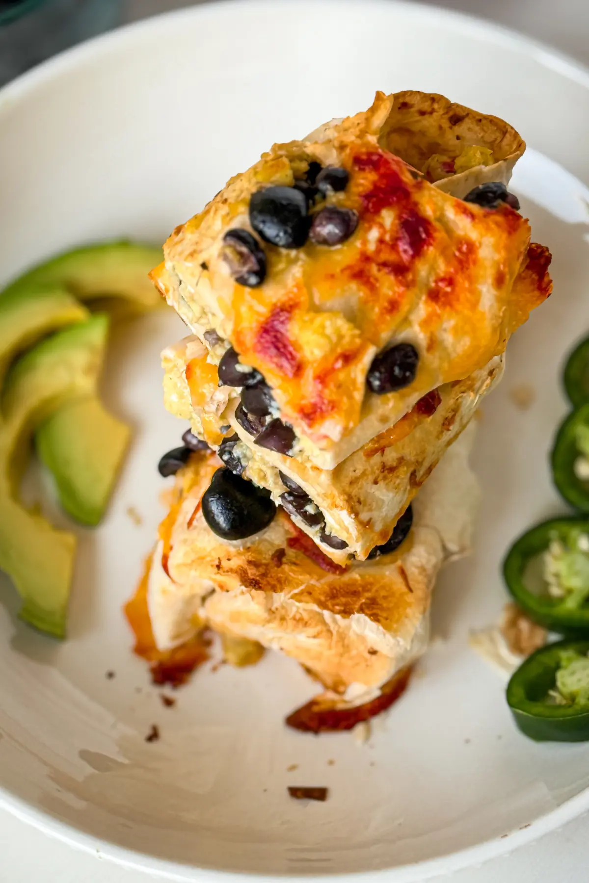 Three halves of a crispy cheese crust high protein breakfast burrito with black beans stacked on top of one another on a plate garnished with avocado slices and jalapenos.