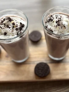 Two Oreo protein shakes in large clear glasses topped with whipped cream and crushed Oreo crumbs.