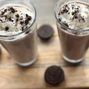 Two Oreo protein shakes in large clear glasses topped with whipped cream and crushed Oreo crumbs.