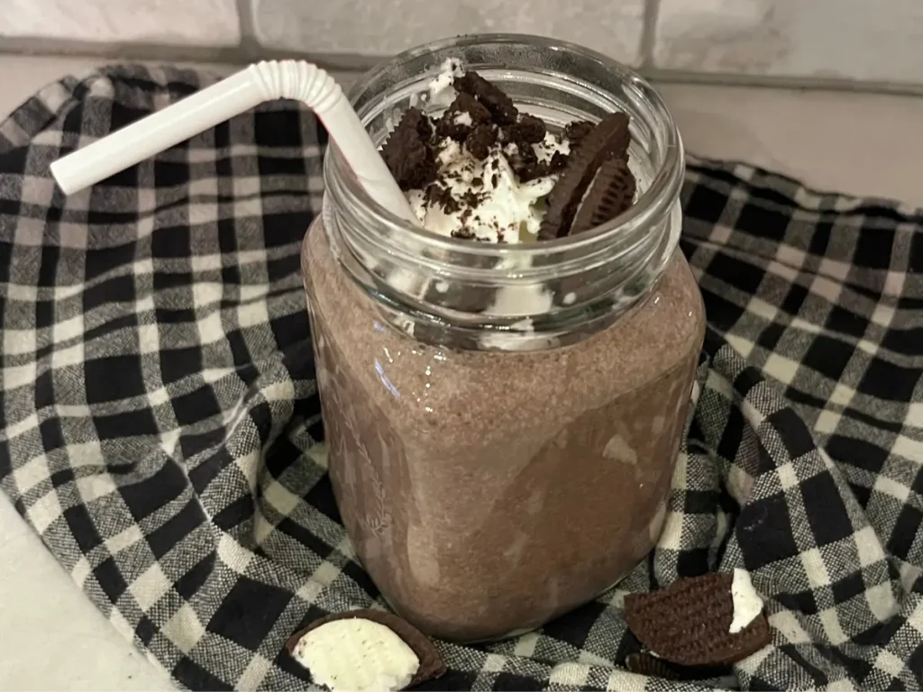 An Oreo protein shake on a black and white towel with a straw in it, topped with whipped cream and Oreo crumbs.
